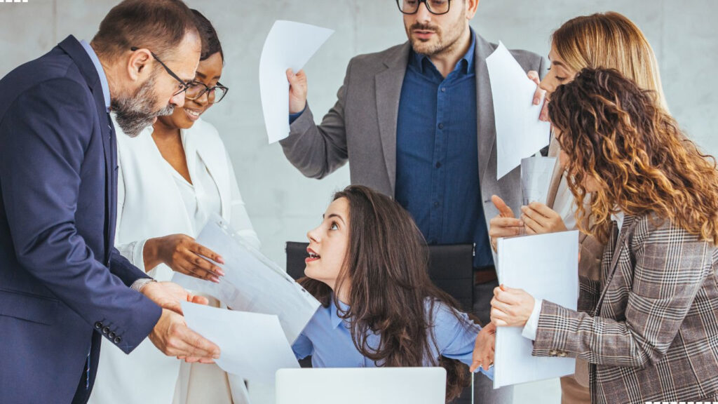 Overwhelmed young female professional surrounded by coworkers with paperwork, representing workplace stress and the importance of soft skills like communication, teamwork, and emotional intelligence. Abhishek Tanwar | xpodcasts